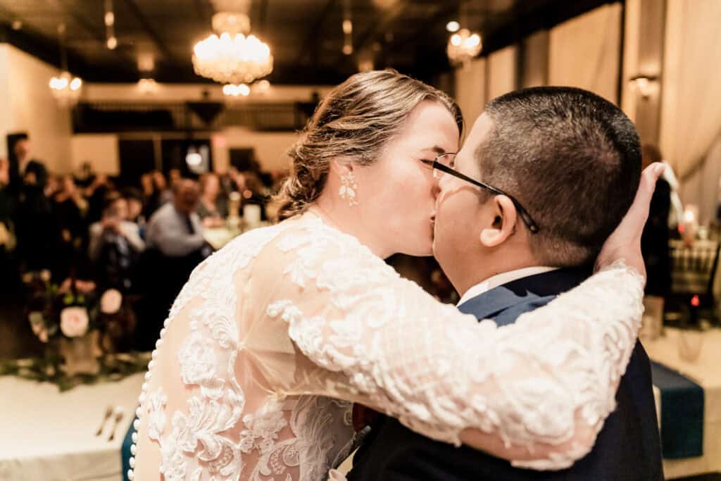 A bride and groom share a kiss at their wedding reception, embraced and surrounded by guests seated at tables in a warmly lit venue.