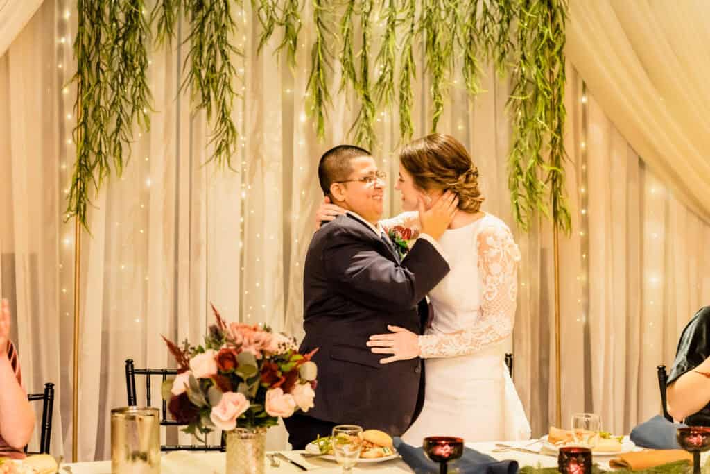 A couple dressed in wedding attire embraces, standing next to a table with plates and floral arrangements, against a backdrop of hanging greenery and string lights.