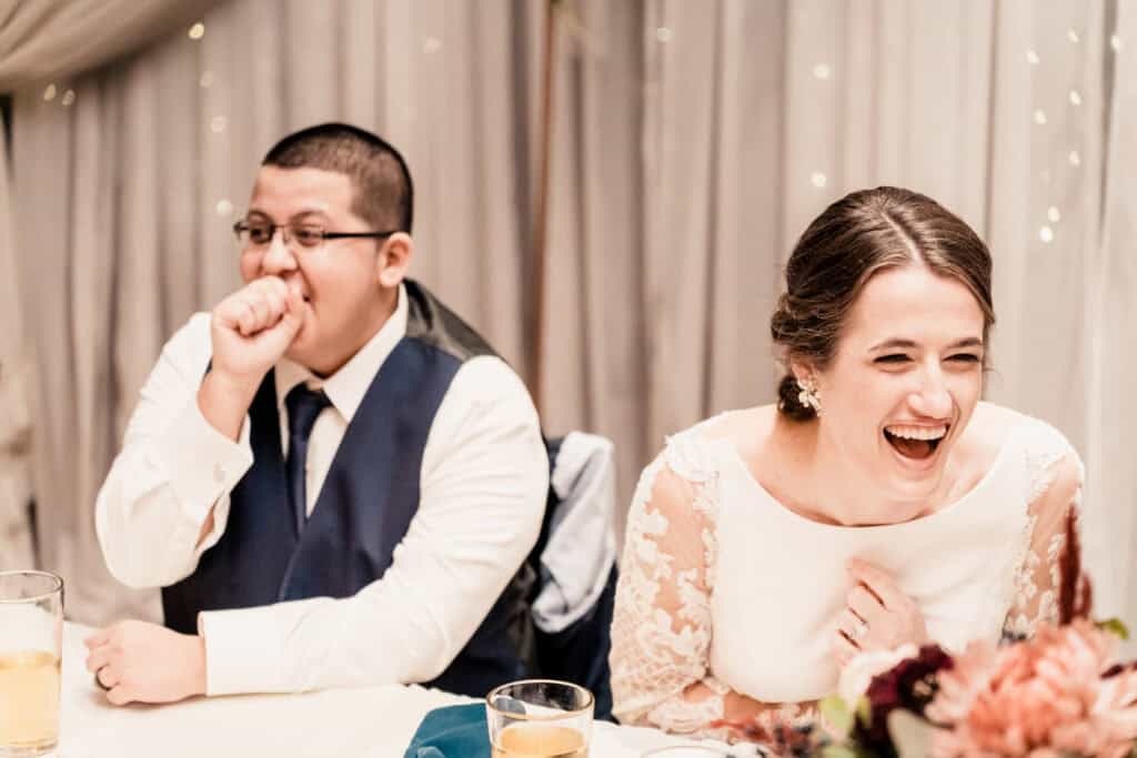 Two people sit at a table, both laughing. The man on the left is dressed in a suit and tie, covering his mouth with his hand, while the woman on the right is in a white dress, laughing openly.