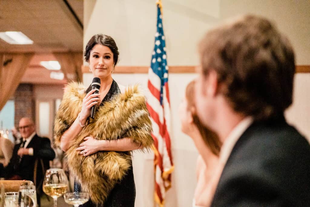 A woman holding a microphone speaks at an indoor event. She is wearing a fur-like shawl, and an American flag is visible in the background. Other seated individuals are in the foreground.