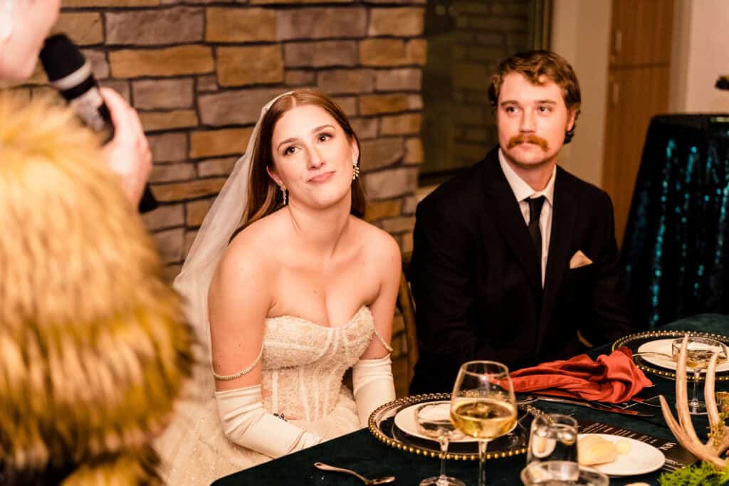 A bride and groom sit at a table listening to a speech. The bride is wearing a white gown and veil, while the groom is in a black suit with a tie. A microphone is visible to the left.