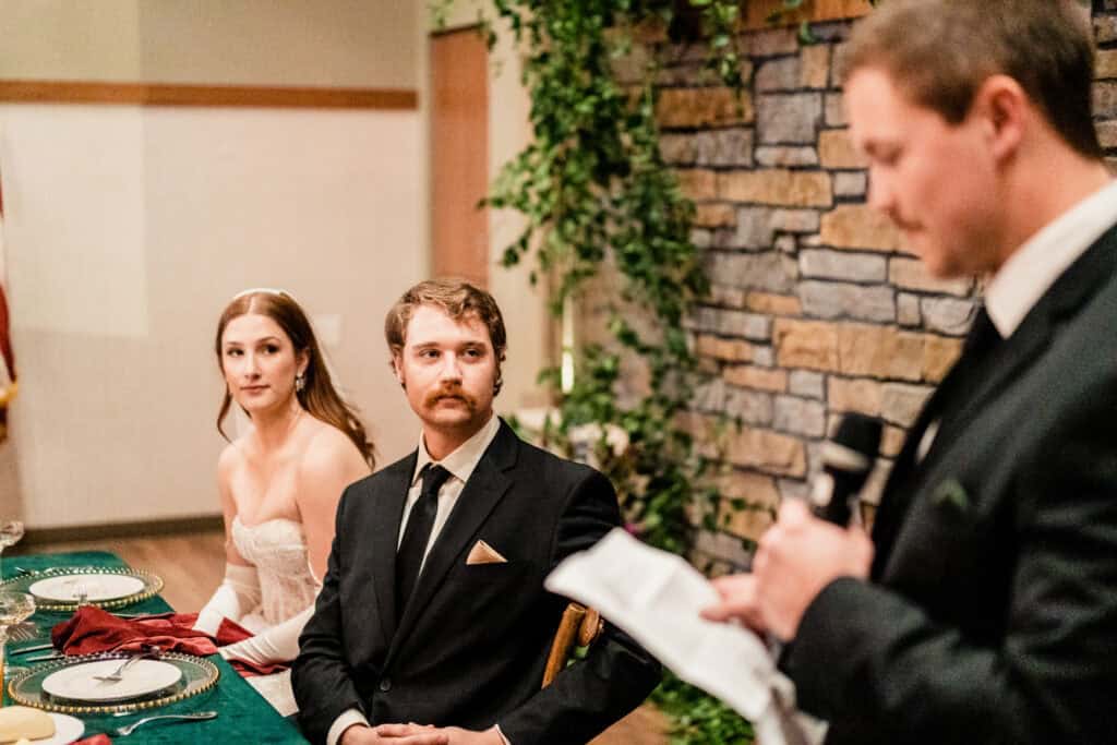 A bride and groom sit at a decorated table, attentively listening to a man in a black suit giving a speech while holding a microphone and a piece of paper.