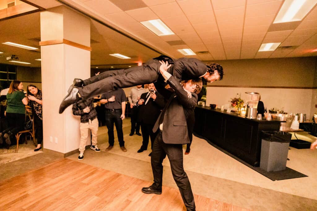 Person in formal attire lifting another person in a playful manner at an indoor social event with onlookers in the background.