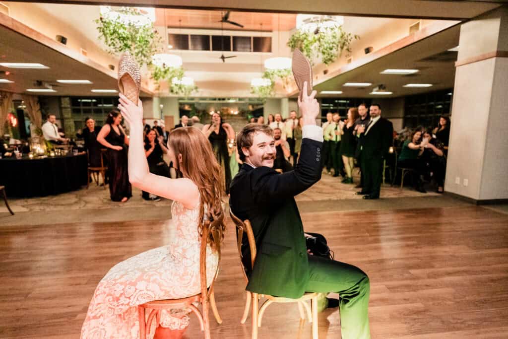 A bride and groom sit back-to-back on chairs holding up shoes during a game at their wedding reception. Guests in formal attire watch and laugh in the background.