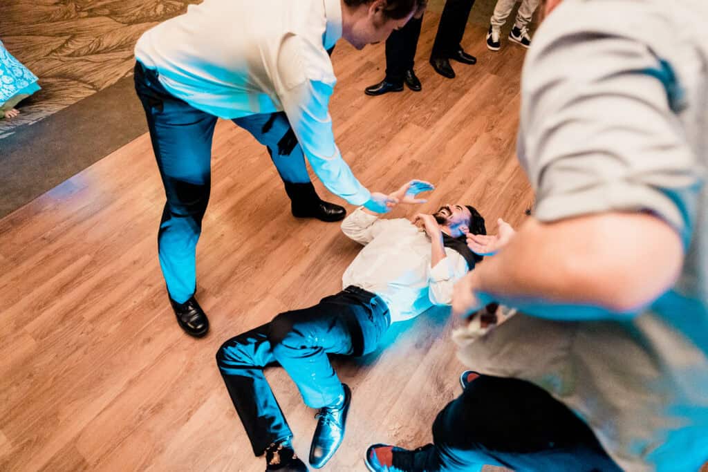 A man lies on the wooden floor while two people around him extend their hands to assist. The scene appears to be in a social gathering or event.