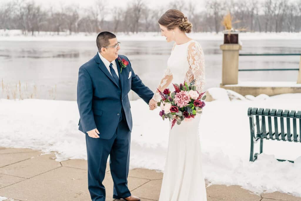 A couple in wedding attire stands outdoors by a snow-covered lakeside. The bride holds a bouquet and the groom, in a suit, looks at her with a smile. A bench and snowy landscape are in the background.