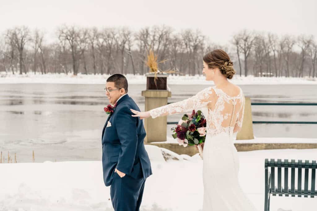 A bride in a white dress and holding a bouquet stands behind a groom in a blue suit, touching his shoulder, near a snowy riverbank with bare trees in the background.