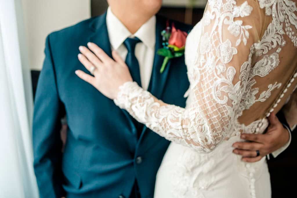 A couple in wedding attire stands close, with the person's hand resting on the other's chest. The bride wears a detailed lace gown.