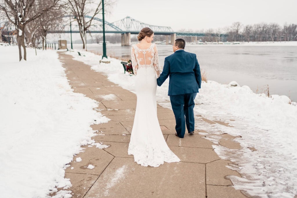 A bride and groom walk hand in hand along a snowy riverside path with a bridge visible in the background.