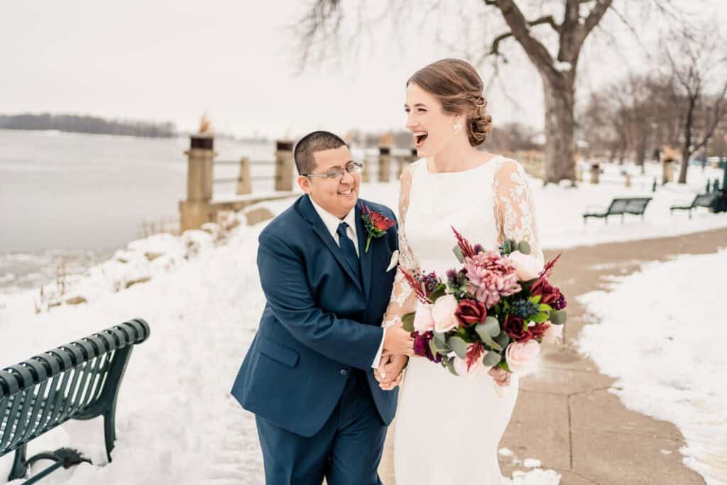 A couple in formal attire shares a joyful moment on a snowy, lakeside pathway. The person in the white dress holds a vibrant bouquet while laughing, as the person in the navy suit smiles.
