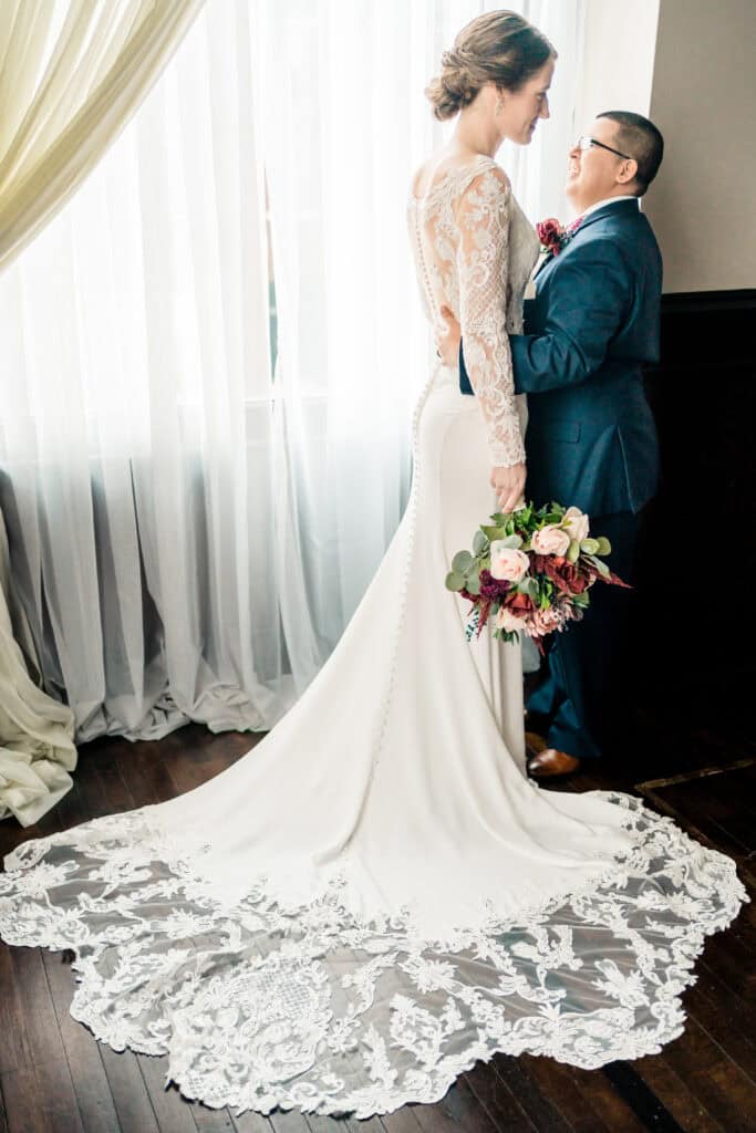 A couple stands by a window; one wears a white lace wedding dress and holds a bouquet, while the other wears a dark suit and glasses, embracing from behind.