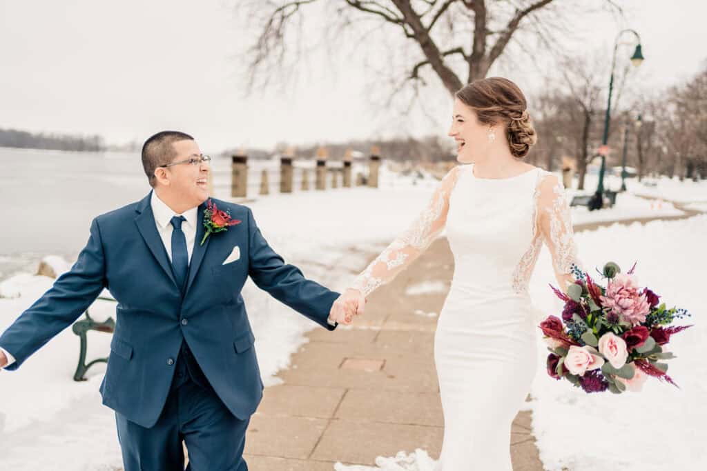 A couple smiles at each other while holding hands and walking on a snowy path. The person on the right wears a white dress, and the person on the left wears a dark suit. The person on the right is holding a bouquet.