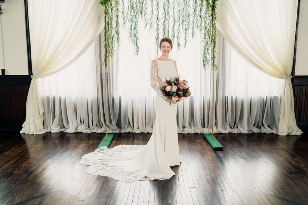 A bride in a white gown with lace sleeves stands indoors on a wooden floor, holding a bouquet of flowers. Draped curtains and greenery hang behind her, creating a decorative backdrop.