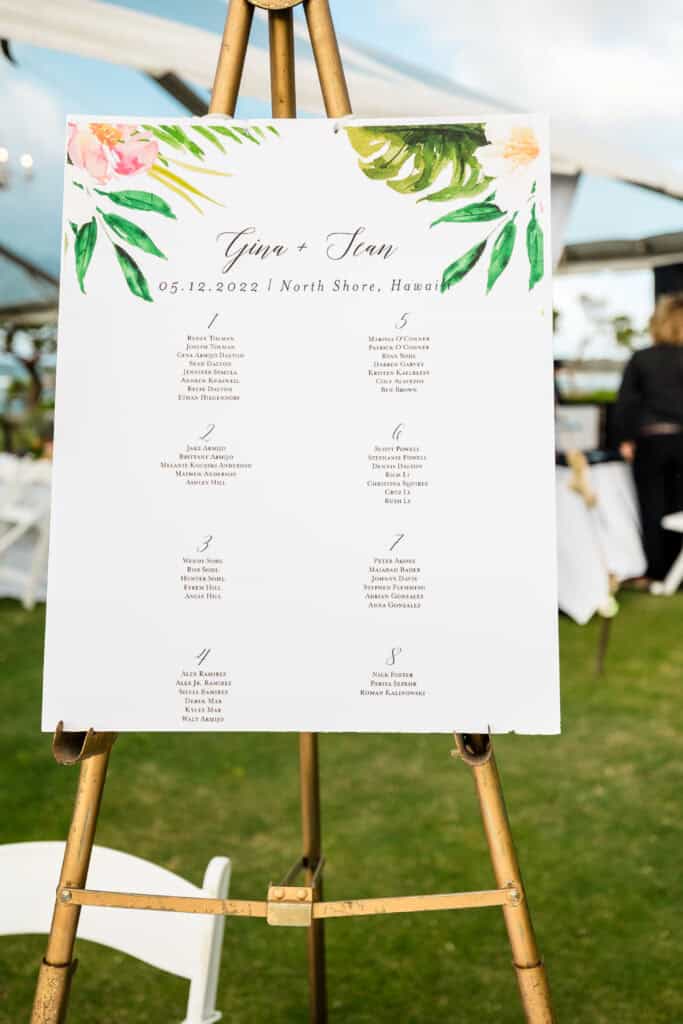 A wedding seating chart on an easel with floral decorations at the top. The chart is titled "Gina & Sean," dated "05.12.2022," and lists table assignments for guests at North Shore, Hawaii.