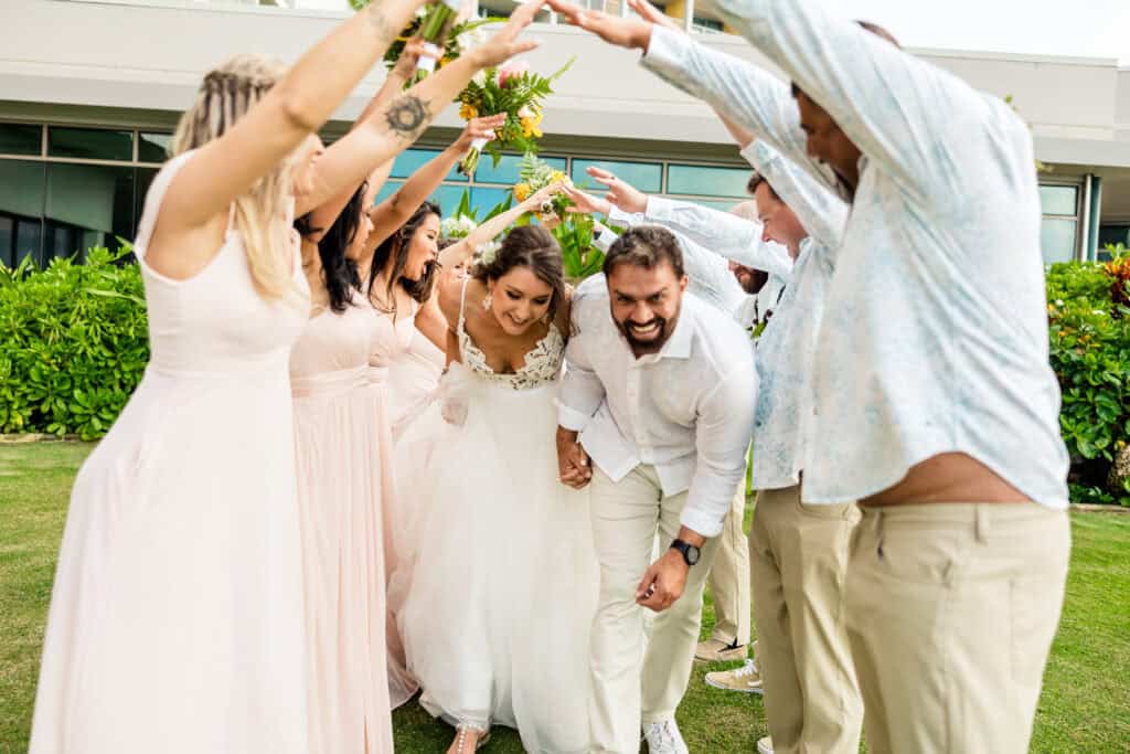 A bride and groom laugh and duck under an arch of raised arms formed by their wedding party, outdoors on a lawn.