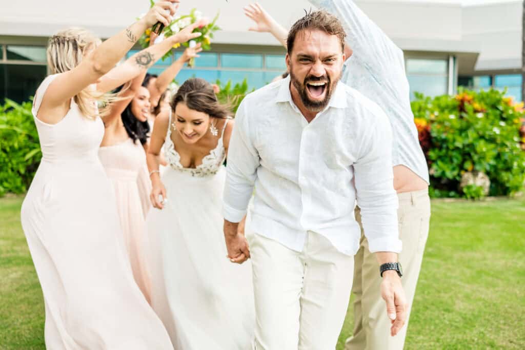 A man and woman in wedding attire, holding hands, walk joyfully under an arch formed by a group of people at an outdoor event with lush greenery in the background.