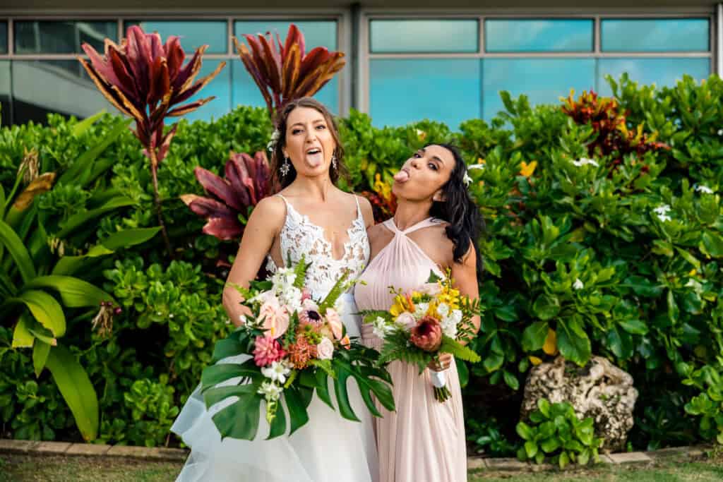 Two women in elegant dresses, holding floral bouquets, make playful faces outdoors in front of lush green foliage and large plants.