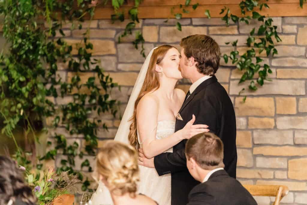 A bride and groom share a kiss in front of a stone wall adorned with greenery. Seated guests are visible in the foreground, observing the couple.