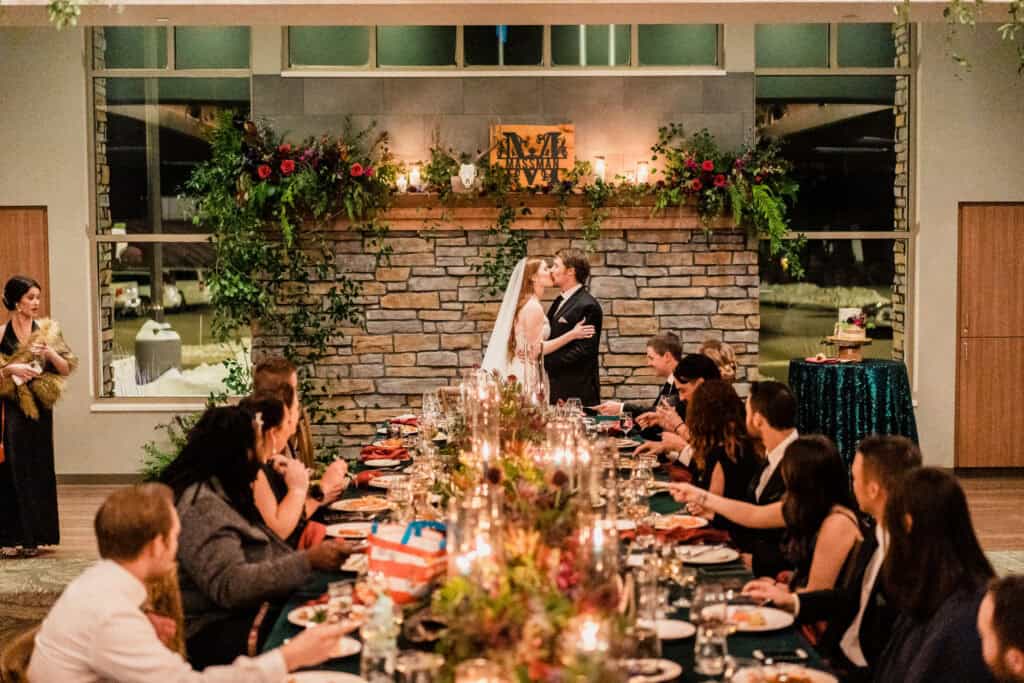 Newlywed couple kisses in front of a stone fireplace while guests seated at a long, decorated table look on during a wedding reception.
