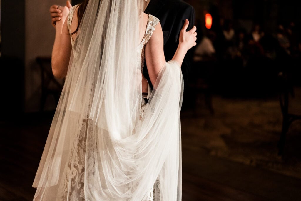 A bride and groom share a dance. The bride's veil flows down her back, and their poses suggest an intimate moment. Scene is dimly lit, creating a warm ambiance.