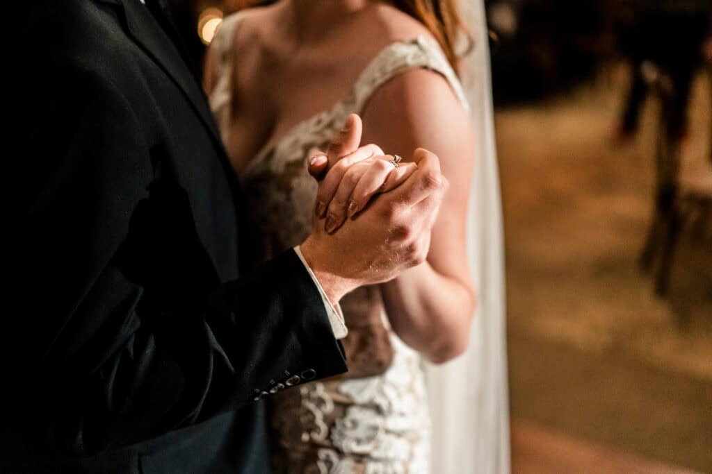 A couple in formal attire holds hands and dances closely, with the woman's arm around the man's neck. The background is slightly out of focus.