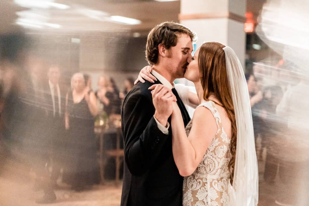 A bride and groom kiss while dancing on their wedding day, with blurred guests in the background.