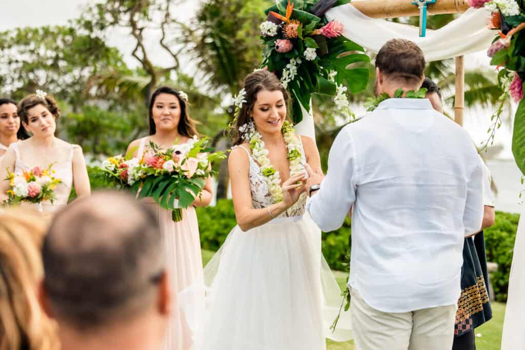 A bride and groom exchange rings in an outdoor ceremony. Bridesmaids with bouquets stand nearby. The setting is decorated with flowers and greenery, with a tropical backdrop.