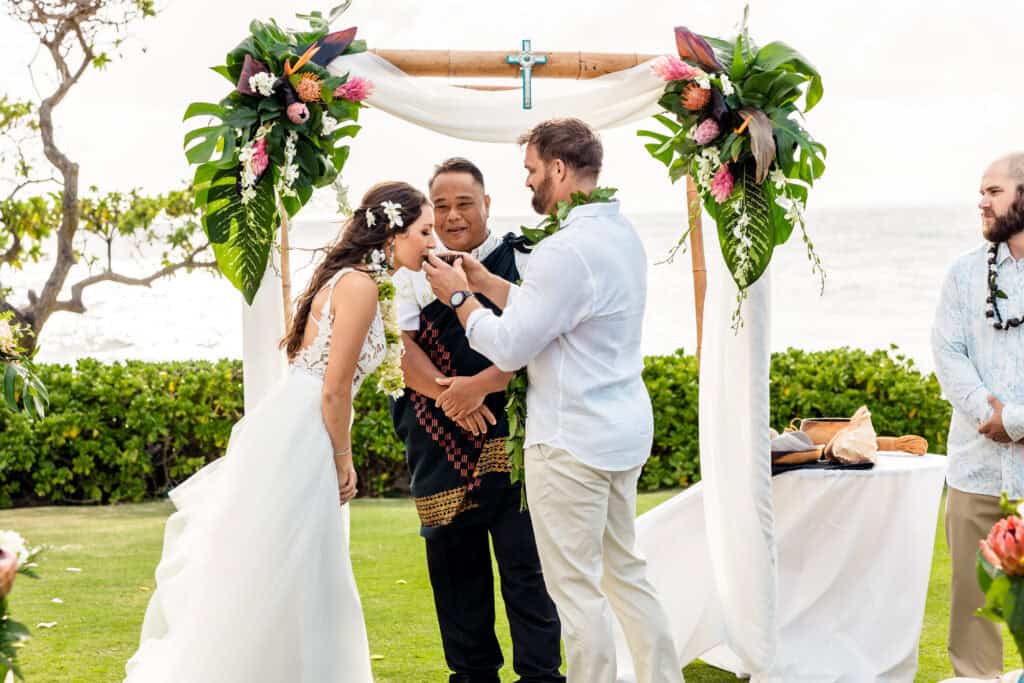 A couple is standing under a decorated wedding arch by the ocean. The man is feeding the woman a piece of food as the officiant looks on. A small table with food is beside them.