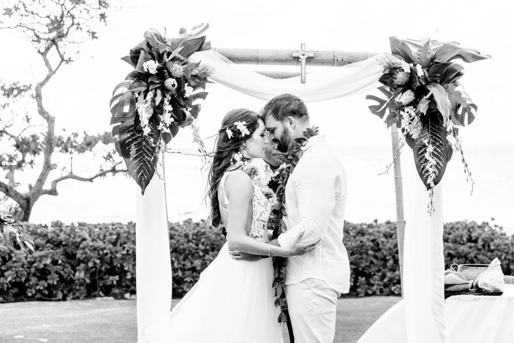 A couple embraces under a decorated wedding arch outdoors, with foliage and ocean in the background. The scene is in black and white.