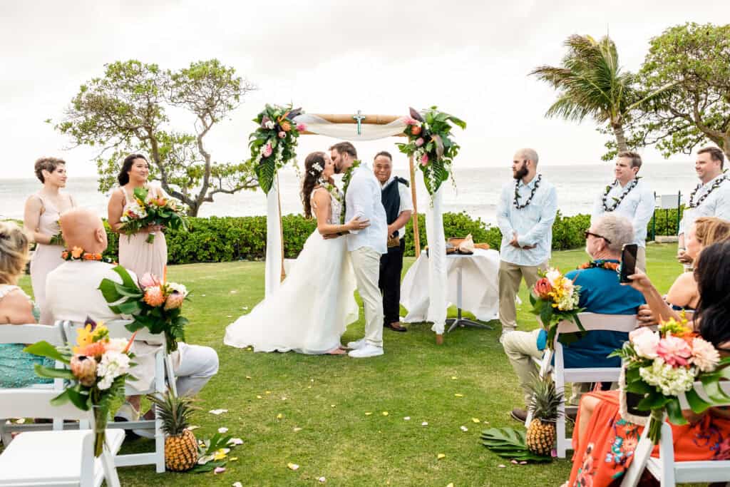 A bride and groom kiss under a decorated arch during an outdoor wedding ceremony by the ocean, surrounded by guests and wedding party members holding bouquets.