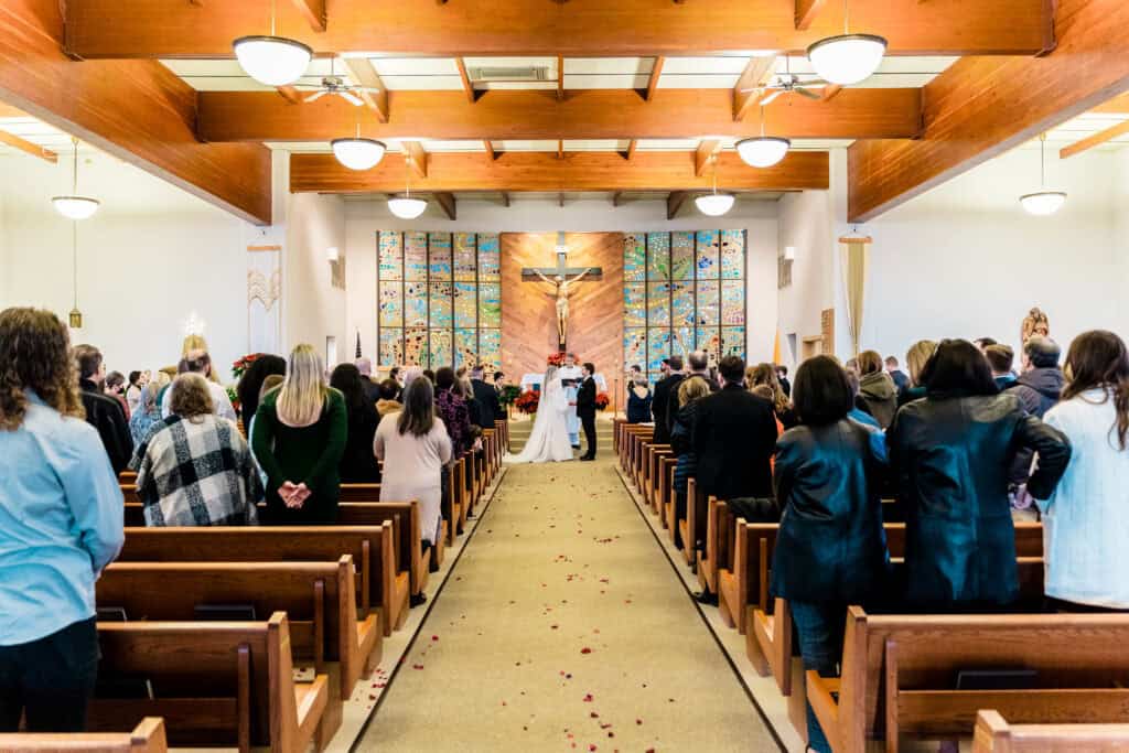 A wedding ceremony is taking place in a church. The couple stands at the altar, while guests are seated on wooden pews. The aisle is decorated with scattered flower petals.