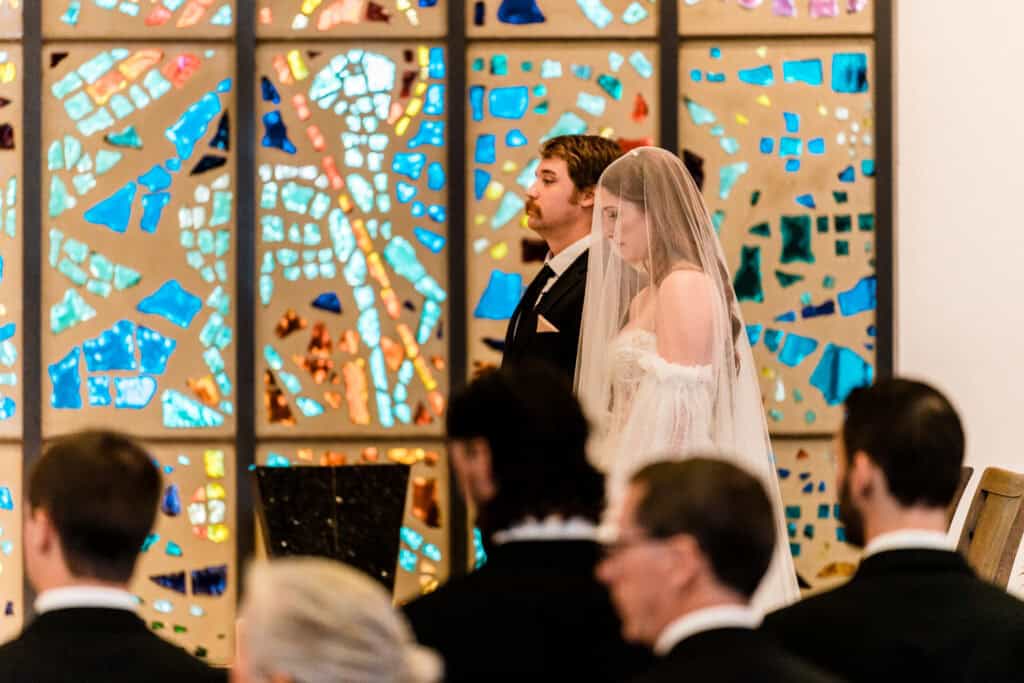 A couple, with the bride veiled, stands at the altar during a wedding ceremony in front of a colorful stained glass window, while guests watch from pews.