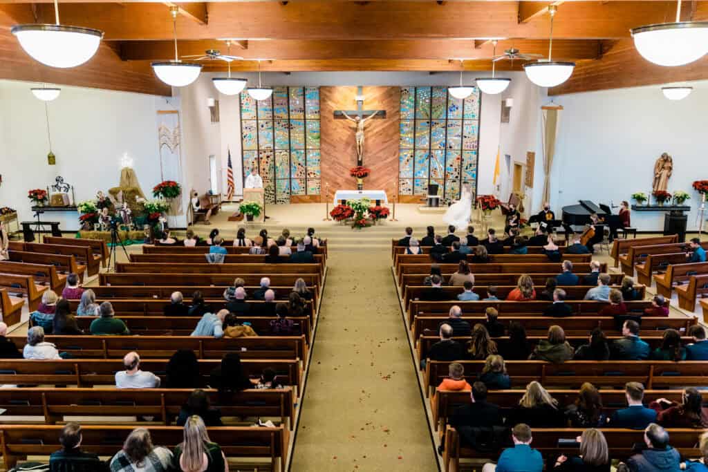 A wide-angle view of a church interior during a wedding ceremony. The congregation is seated in wooden pews, and a priest stands at the altar. Stained glass windows and a crucifix are visible behind the altar.