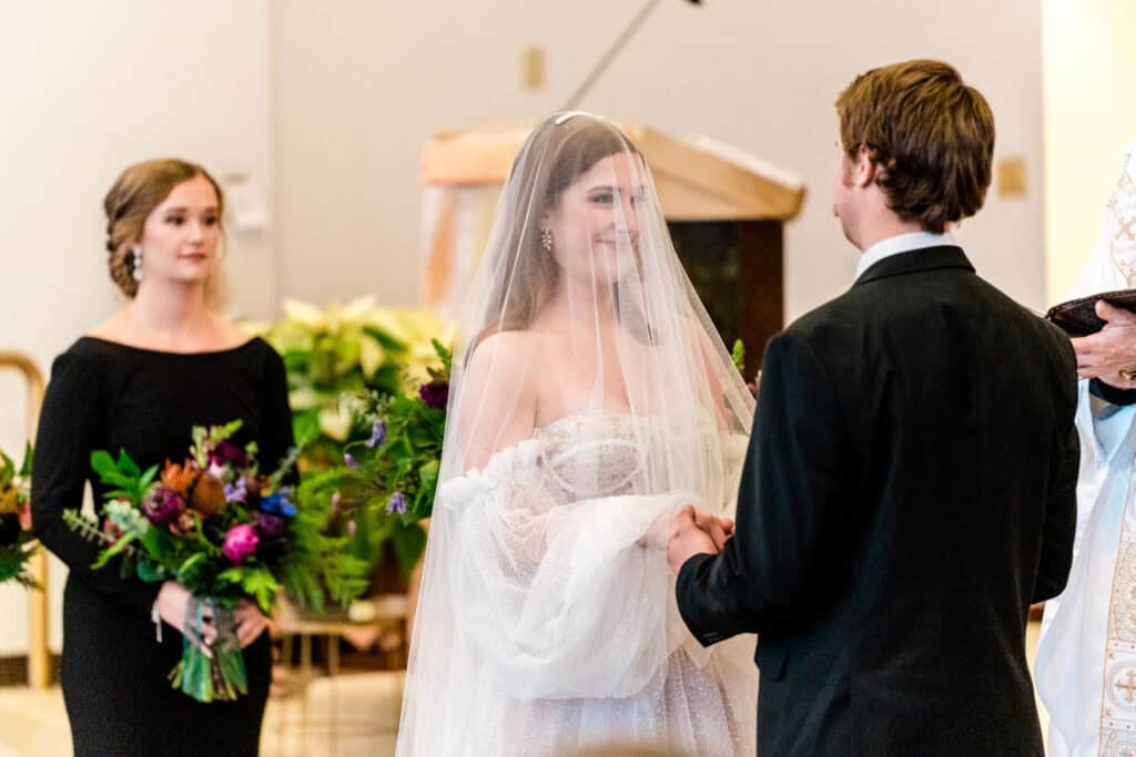 A bride and groom stand facing each other during a wedding ceremony. The bride is wearing a white dress and veil, while the groom is in a black suit. A bridesmaid holding a bouquet stands nearby.
