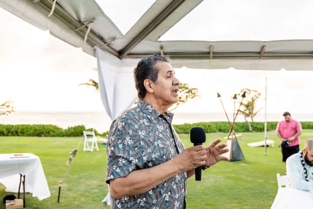 A man in a patterned shirt holds a microphone and speaks outdoors under a canopy tent, with a grassy area and the ocean in the background.