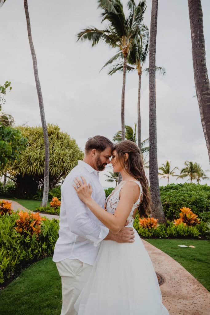 A couple embraces tenderly outdoors, surrounded by tropical plants and palm trees, both wearing white attire.