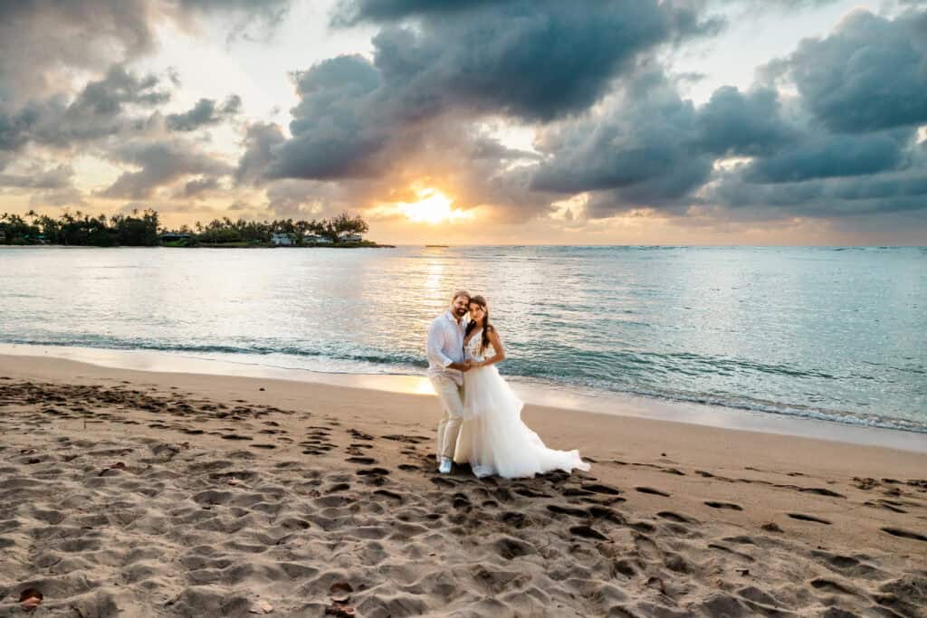 A couple in wedding attire embraces on a beach at sunset, with footprints in the sand and dramatic clouds in the sky.