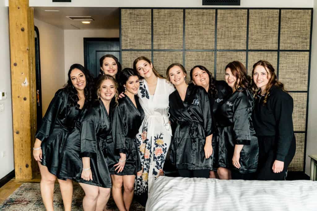 A bride in a white robe stands with eight women in black robes, all smiling and posing together in a room with a tiled wall.