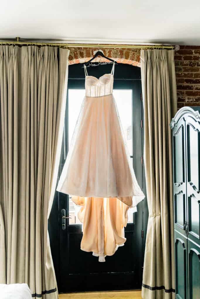 A white wedding dress hangs on a black hanger in front of a large window with beige curtains, displaying its full length and detailed design.
