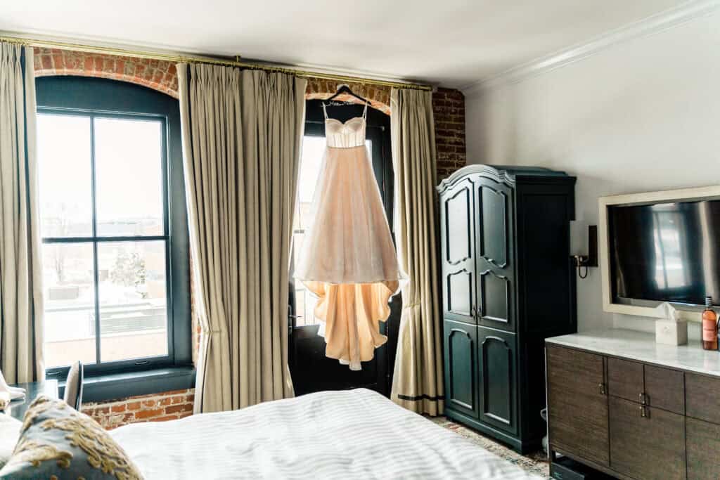 A wedding dress hangs on a black hanger in front of a window with beige curtains in a well-lit bedroom with brick walls, an armoire, a bed, a TV, and a dressing table.
