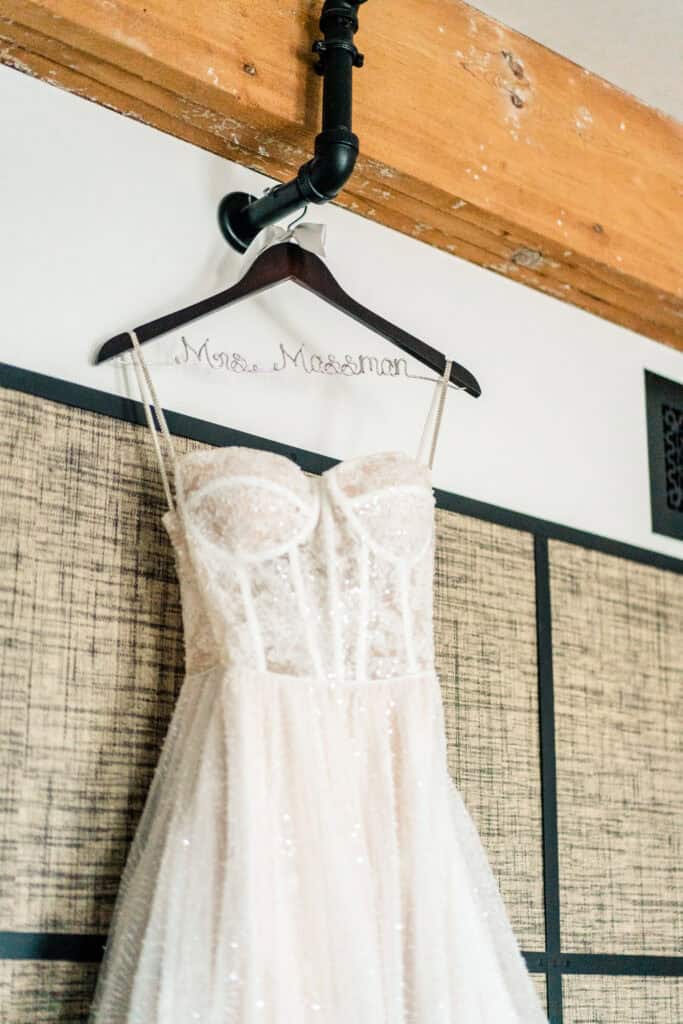 A white, strapless wedding dress with a sequined bodice hangs on a black hanger with "Mrs. Mossman" written in wire. The background features wooden beams and a checkered wall.