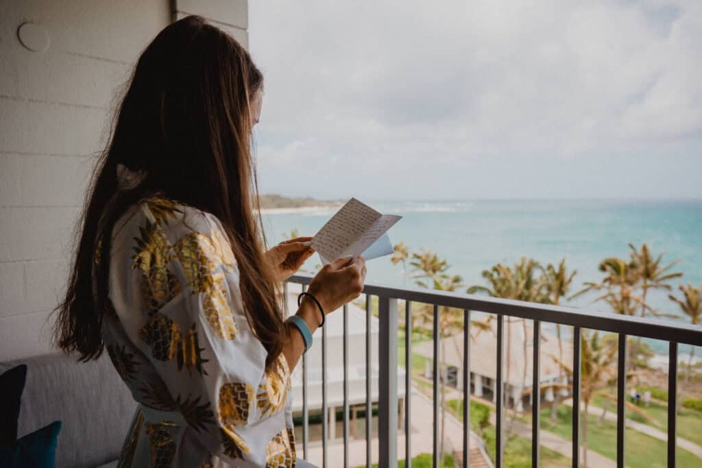 A person with long hair reads a handwritten card while standing on a balcony overlooking a beach scene with palm trees and ocean in the background.