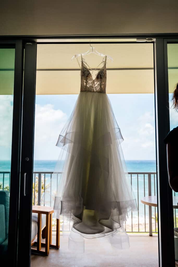 A white wedding dress with a tiered tulle skirt and lace bodice hangs on a hanger in front of an open glass door overlooking an ocean view.