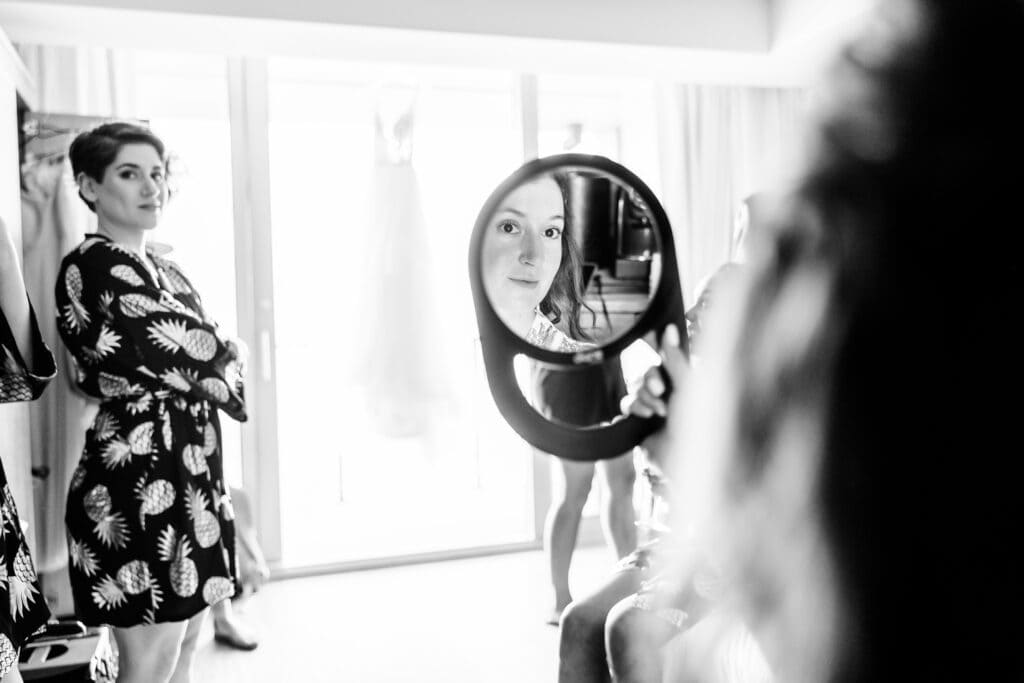 A woman stands with her arms crossed while another woman holds up a mirror in which her face is reflected. A dress hangs in the background. The photo is in black and white.