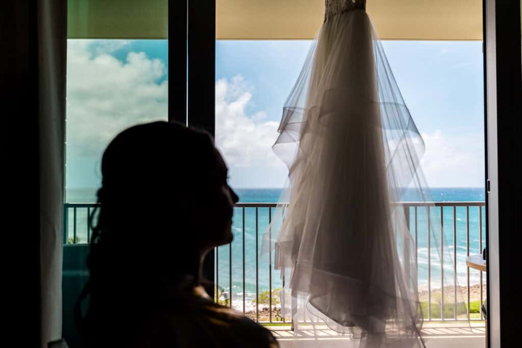 Silhouette of a person facing a hanging wedding dress in front of a balcony with a view of the ocean.