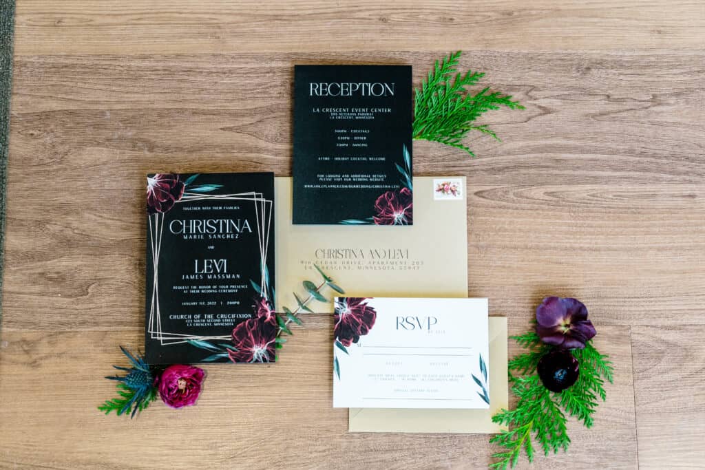 Wedding invitation suite with cards for ceremony, reception, and RSVP placed on a wooden surface, accentuated by green leaves and purple flowers.