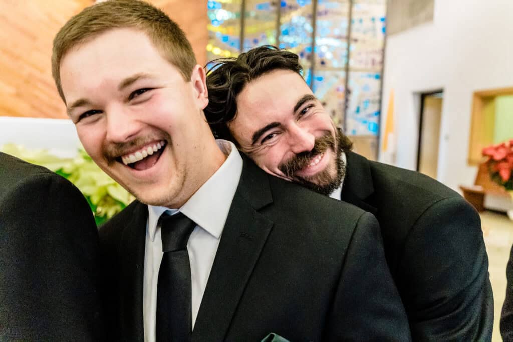 Two men in suits are smiling and embracing each other. One man is standing in front while the other man hugs him from behind. They are indoors with stained glass windows in the background.