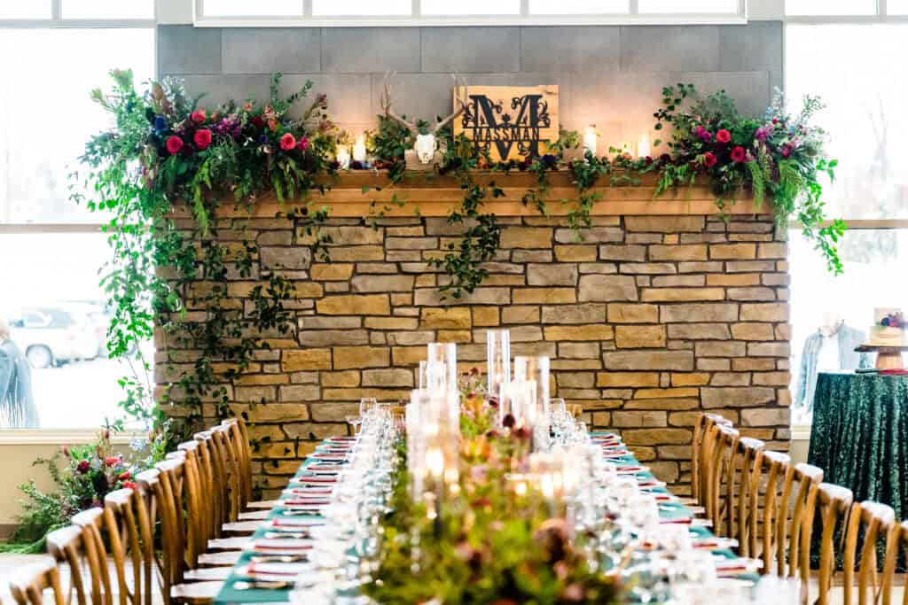 A dining table set for an event is adorned with tableware and greenery. In the background is a stone fireplace decorated with flowers, green foliage, and a wooden sign with the name "Masseman.