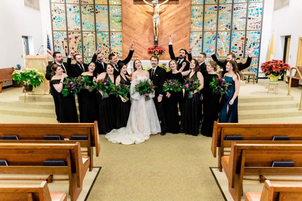 A bride and groom stand with their wedding party in a church. They are all smiling and raising their hands, and some hold bouquets.