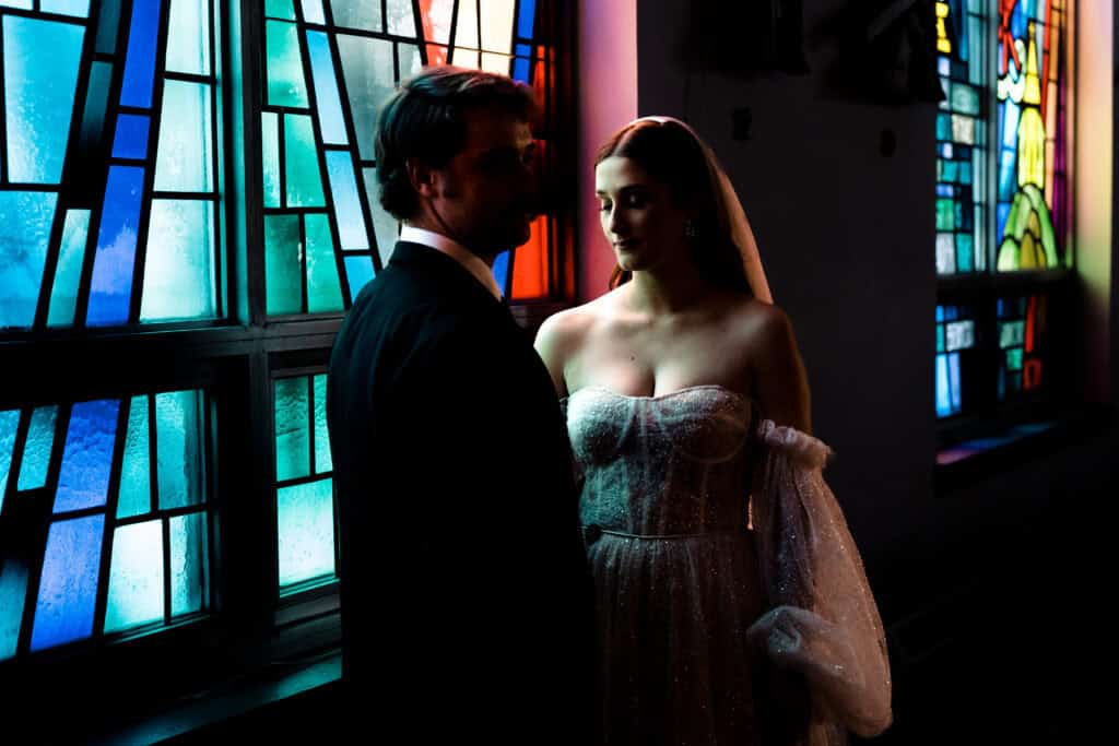 A couple, dressed in wedding attire, stands near colorful stained glass windows. The bride is in a gown with a veil and the groom is in a black suit. They are facing each other in a dimly lit setting.